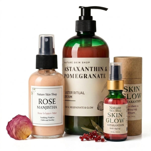 A skincare gift set from Nature Skin Shop on a white background, featuring a Rose Manjistha mist, a large Astaxanthin & Pomegranate cream bottle, a Skin Glow astaxanthin spray, a brown paper tube, and decorative rose petals and pomegranate seeds.