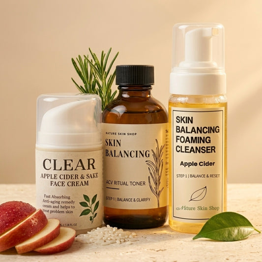 Three skincare products arranged on a light stone surface with sliced apple and a green leaf as props: a white face cream tube labeled Clear, an amber bottle labeled Skin Balancing Facial Toner, and a yellow cleansing bottle labeled Skin Balancing Foaming Cleanser. The products are shown in a clean, styled studio setup with warm beige background.