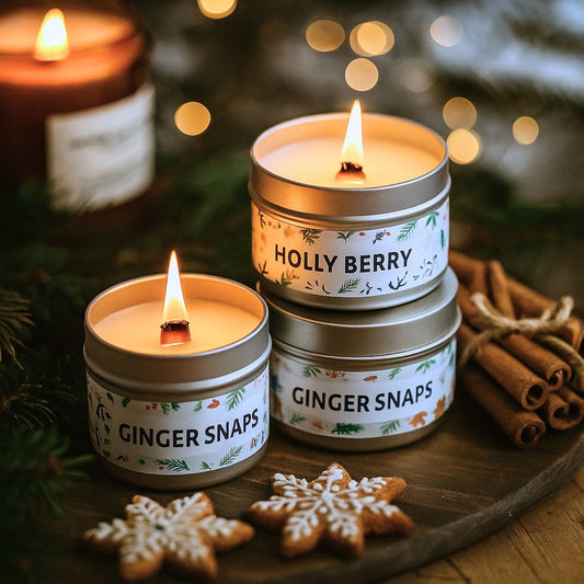 Three small candles in tins labeled 'Holly Berry', 'Ginger Snaps', and 'Ginger Snaps' on a wooden surface with Christmas decorations.