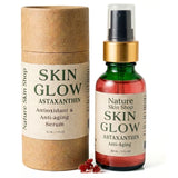 A bottle of Nature Skin Shop Skin Glow Astaxanthin anti-aging serum next to its brown kraft paper packaging cylinder, with a few red astaxanthin crystals in front, on a white background.