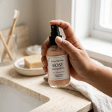 Hand holding a bottle labeled 'Rose Manjistha' on a bathroom counter.