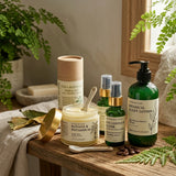A curated collection of botanical skincare products arranged on a rustic wooden table with a linen cloth and green ferns in the background. The products include a green pump bottle of 'Botanical Accept Lotion', a green spray bottle of 'N.