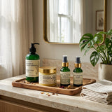 A set of botanical skincare products arranged on a wooden tray on a marble bathroom counter, with a mirror, curtains, and a potted plant in the background.