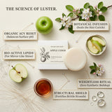 An Apple Cider Shine Ritual Bar by Nature Skin Shop is displayed in a styled flat-lay with labeled callouts explaining its ingredients: organic ACV reset, bio-active lipids, botanical infusion, weightless ritual, and structural shield. The bar and its white box are centered, surrounded by a green apple, apple slices, white blossoms, and a glass of amber liquid on a light beige textured background.