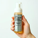 Hand holding a bottle of 'Skin Balancing Foaming Cleanser' with apple cider vinegar on a light background