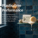 A boxed apple cider hair care product sits on a glass shelf against a dark blue tiled wall, with a bar of soap and a small bowl nearby. Large text on the left reads 'Plastic-Free Performance' and 'High-performance hair care that delivers professional results without the plastic waste.