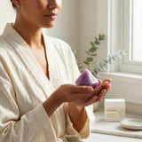 Woman in a white robe holding a purple crystal in a bright room with a plant and box in the background.