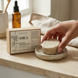 Person holding a natural sponge next to an Amla product box on a wooden surface.