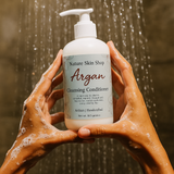 Person holding a bottle of 'Nature Skin Shop Argan Cleansing Conditioner' under running water.