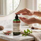 A bottle of Nature Skin Shop Astaxanthin & Pomegranate Ritual Cream sits on a marble countertop with hands reaching toward the pump, in a bright bathroom setting.