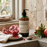 A pump bottle of Nature's Skin Drop Mastaxanthin & Pomegranate Claster Ritual Cream stands on a stone countertop beside pomegranate halves, a cloth, and greenery in a kitchen-like setting.