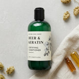 Green bottle of 'Beer & Keratin' conditioner on a light background with dried flowers and a towel.