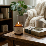Candle labeled 'Black Sea' on a wooden table with a mug, book, and glasses in a cozy room.