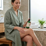 A woman in an olive green robe sits on a wooden stool in a bathroom, applying a glossy body cream from a green pump bottle to her leg.