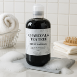 Bottle of Charcoal & Tea Tree Detox Bath Gel on a bathroom counter with towels and a brush in the background.