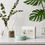 Green soap bar and box on a marble surface with plants and jars in the background