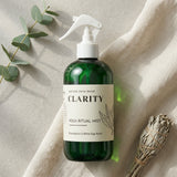 Green bottle labeled 'Clarity' with a white label on a beige fabric background with eucalyptus leaves and sage.