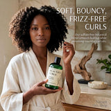 A woman in a cream robe holds a green bottle of Nature Skin Shop Beautiful Curls Co-washed Shampoo in a bathroom setting. Text on the image reads 'SOFT, BOUNCY, FRIZZ-FREE CURLS'.