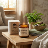 Earth & Oakmoss candle on a wooden table with books and a plant in the background