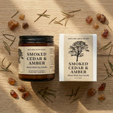 Smoked Cedar & Amber candle and packaging on a wooden surface with natural elements.