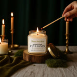Candle being lit with a 'Nature Skin Shop' candle labeled 'Full Moon' on a wooden stand.