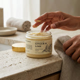 Jar of 'Ginger & Lime' body scrub with a hand reaching towards it on a stone surface.