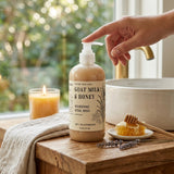 A bottle of Nature Skin Shop Goat Milk & Honey Nourishing Ritual Wash sits on a rustic wooden counter next to a lit candle, a white bowl, and a hand reaching toward the pump dispenser.