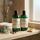 Two green bottles of Nature Skin Shop Gugo & Caffeine hair care products, including strengthening shampoo and strengthening conditioner, displayed on a stone bathroom counter with a folded towel, eucalyptus leaves, coffee beans, and a wooden comb nearby.