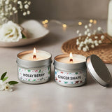 Two lit candles in metal tins labeled 'Holly Berry' and 'Ginger Snaps' on a decorative surface with flowers.