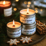 Three small candles in tins labeled 'Holly Berry', 'Ginger Snaps', and 'Ginger Snaps' on a wooden surface with Christmas decorations.