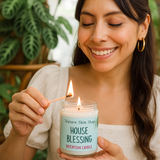 Woman lighting a candle labeled 'House Blessing' from Nature Skin Shop with plants in the background.