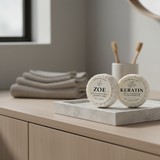 Two soap bars labeled 'ZOE' and 'Keratin' on a bathroom counter with towels and toothbrushes in the background.
