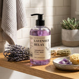 Lavender Relax Therapeutic Ritual Wash by Nature Skin Shop in a clear pump bottle, styled on a wooden shelf with dried lavender, a towel, a small plant, and bath stones in a bright bathroom setting.