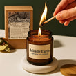 Person lighting a 'Middle Earth' artisan soy candle with a match, on a white surface with a green background.