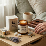 Candle labeled 'Moon Stone' on a wooden surface with a cup of tea, books, and lavender.