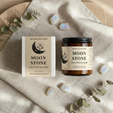 Moon Stone candle jar and packaging on a textured surface with eucalyptus leaves and crystals.