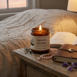 Candle labeled 'Moon Stone' on a wooden surface with a book and crystals in a bedroom setting.