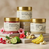 Three jars of natural skin care products with ingredients like bananas, ginger, and limes on a wooden surface.