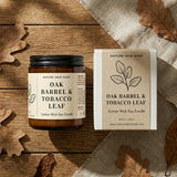 Candle jar and packaging of 'Oak Barrel & Tobacco Leaf' candle from Nature Skin Shop on a wooden surface with leaves.