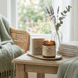 Candle labeled 'The Purifying Ritual' on a wooden table with books and a vase of lavender in the background.