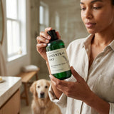 Woman holding a bottle of 'Ancestral Oat' pet wash with a dog in the background.