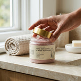 Hand opening a jar of Pink Himalayan and Rose Manjistha scrub on a bathroom counter.