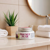 Jar of Prickly Pear body cream on a bathroom counter with towels and a plant in the background.