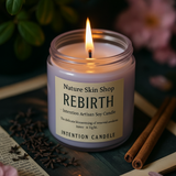 Candle labeled 'Rebirth' from Nature Skin Shop on a wooden surface with cinnamon sticks and flowers.