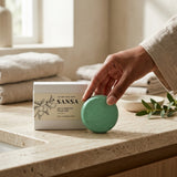 A hand holds a round mint green soap bar next to its white SANSA box on a beige stone countertop in a softly lit bathroom setting.