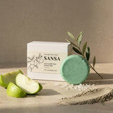 SANSA Acy Clarifying Ritual Bar soap shown with its white box packaging, alongside a mint green round bar of soap, green apple slices, and a sprig of olive leaves on a textured beige surface.
