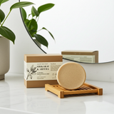Bar of soap on a wooden soap dish with packaging in the background, on a white surface with a plant.