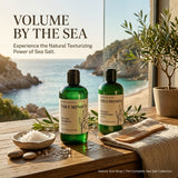 Two green bottles of Volumnous hair care products, Sea Salt Shampoo and Seaweed Conditioner, displayed on a wooden table with a beach background. The scene includes a small bowl of sea salt, a wooden brush, and a potted olive tree.