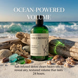 A green bottle of VOLUMINOUS Sea Salt Shampoo from Nature's Skin Shop is displayed on a sandy beach with driftwood, seashells, and colorful sea glass. The background shows ocean waves under a blue sky, with promotional text overlays reading 'OCEAN-POWERED VOLUME' and 'Salt-infused detox clears heavy oils to reveal airy, textured volume that lasts 24 hours.